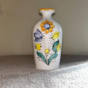 Puede incluir: Jarrón de cerámica blanca con diseño floral. El jarrón tiene un borde amarillo y está decorado con flores amarillas, azules y verdes. Las flores están dispuestas en forma de ramo, con hojas y tallos verdes.