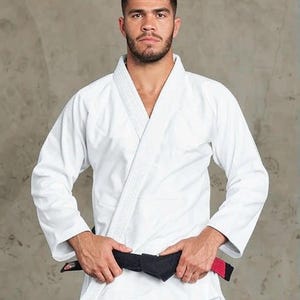 May include: A white martial arts gi featuring a thick, textured lapel and long sleeves. A black belt with a red stripe detail is tied around the waist of the garment. The fabric appears durable and structured, designed for grappling sports.