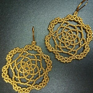 May include: A pair of gold-colored beaded earrings. Each earring features a floral design with a delicate, openwork pattern. The earrings have a hook closure and are displayed against a dark background.