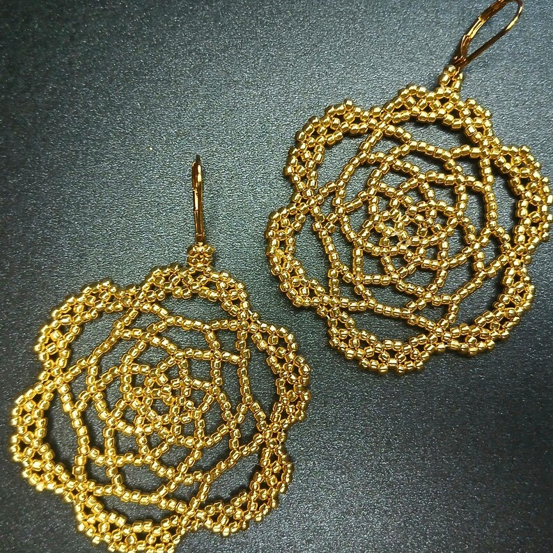 May include: A pair of gold-colored beaded earrings. Each earring features a floral design with a delicate, openwork pattern. The earrings have a hook closure and are displayed against a dark background.