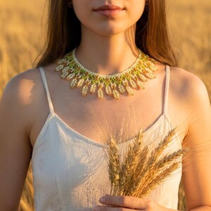 May include: A beaded necklace with white, green, and gold accents. The necklace is worn over a cream-colored dress. The image also features a bundle of wheat stalks, suggesting a natural or bohemian theme. The necklace's design is intricate and detailed.