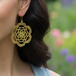 May include: Gold-colored filigree earrings in a floral design. The earrings are a large, openwork style with a detailed, layered pattern. The earrings hang from a simple gold hook. The background is a soft focus of green and purple.