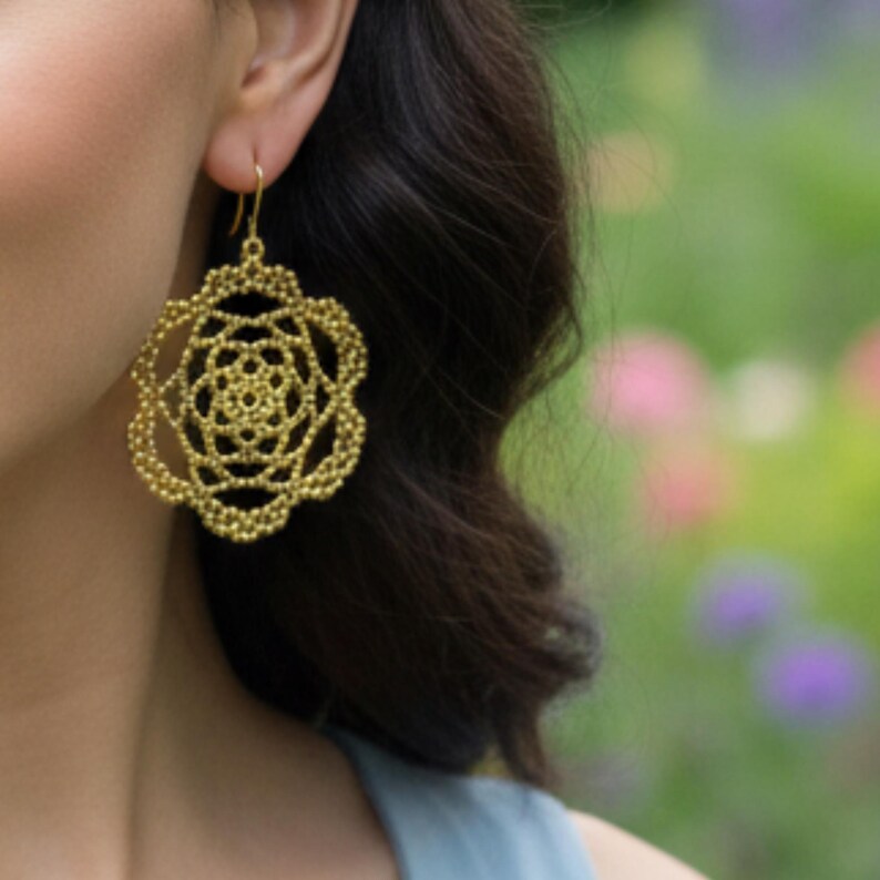 May include: Gold-colored filigree earrings in a floral design. The earrings are a large, openwork style with a detailed, layered pattern. The earrings hang from a simple gold hook. The background is a soft focus of green and purple.