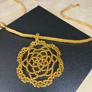 May include: A gold-colored necklace with a beaded pendant in a floral design. The pendant is a detailed openwork pattern, and the chain is also made of small, gold-colored beads. The necklace is displayed on a dark surface.