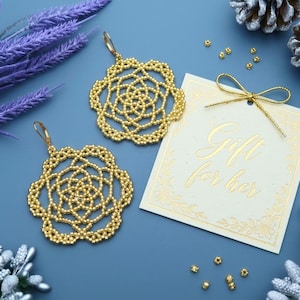 May include: A pair of gold beaded earrings with a floral design. The earrings are displayed with a gift tag that reads "Gift for her" on a blue surface. Decorative elements include sprigs and pinecones.