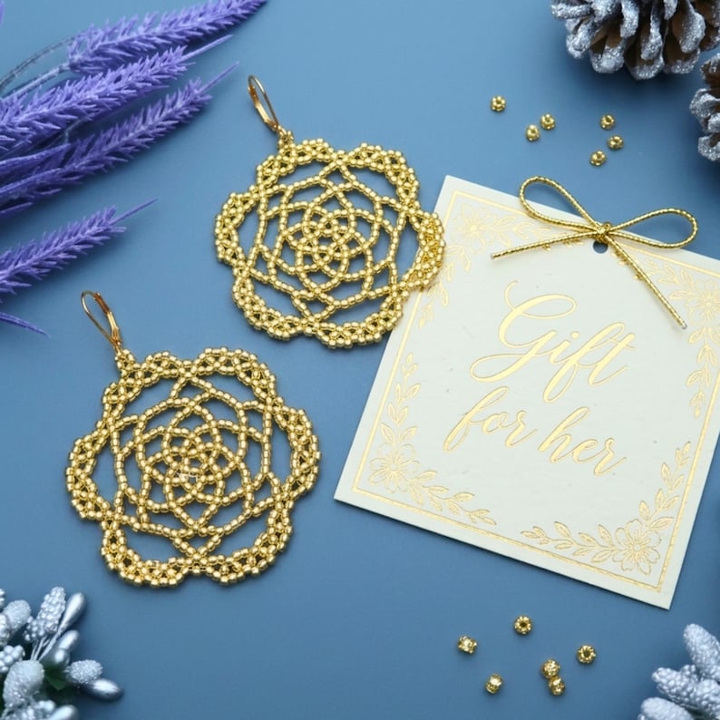 May include: A pair of gold beaded earrings with a floral design. The earrings are displayed with a gift tag that reads "Gift for her" on a blue surface. Decorative elements include sprigs and pinecones.