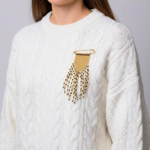 Puede incluir: Un broche de imperdible en tono dorado que presenta una sección de malla tejida triangular en la parte superior y una serie de hilos de flecos de cuentas colgantes. Los flecos consisten en pequeñas cuentas negras intercaladas con diminutos acentos metálicos dorados.
