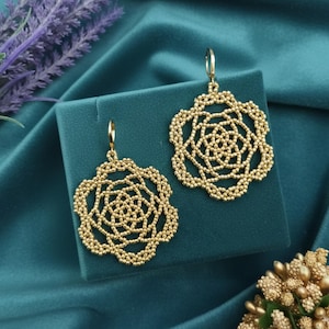 May include: A pair of gold-colored, openwork earrings with a floral design. The earrings are displayed on a teal-colored surface, with a teal background. The earrings have a detailed, beaded appearance and a hook closure.
