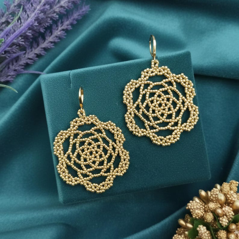 May include: A pair of gold-colored, openwork earrings with a floral design. The earrings are displayed on a teal-colored surface, with a teal background. The earrings have a detailed, beaded appearance and a hook closure.