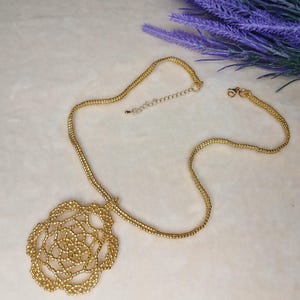 May include: A gold-colored necklace with a floral pendant. The pendant is a detailed openwork design, and the chain is a delicate beaded style. The necklace is displayed on a light-colored surface with purple flowers in the background.