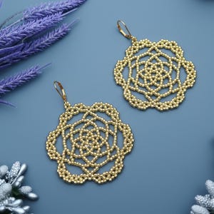 May include: A pair of gold-colored beaded earrings. Each earring features a floral design with intricate detailing. The earrings have a lever-back closure and are displayed on a blue background.