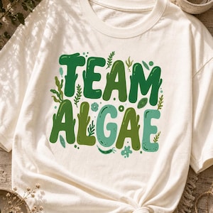Peut inclure: Un t-shirt à manches courtes blanc cassé avec le texte "TEAM ALGAE" en lettres vertes grasses et arrondies. Le motif est décoré de diverses petites illustrations botaniques vertes d'algues, de plantes aquatiques et de points autour du texte.