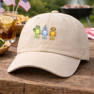 May include: A beige baseball cap with three cartoon robots in yellow, blue, and yellow. The robots have the numbers 01, 10, and 11 on their chests. The cap has a curved peak and a fabric strap.