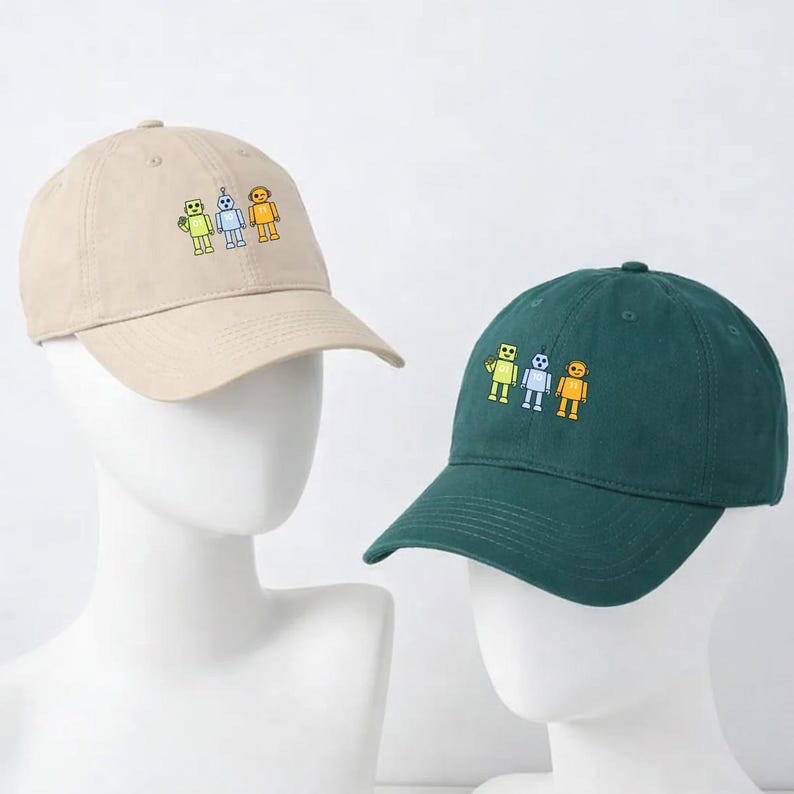 May include: Two baseball caps, one beige and one dark green, each featuring an embroidered design of three colourful robots. The caps have a curved peak and are displayed on mannequin heads. The robots are yellow, blue, and orange.