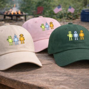 May include: Three baseball caps in beige, pink, and dark green, each embroidered with a robot design. The robots are yellow, blue, and orange, with the numbers 01, 10, and 11 on their chests. The caps are on a wooden surface.
