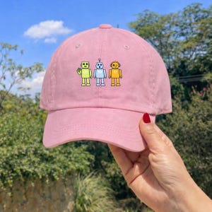 May include: A pink baseball cap with an embroidered design of three robots. The robots are in yellow, blue, and green, each with a unique expression and number. The cap is made of a soft fabric and has a curved peak.