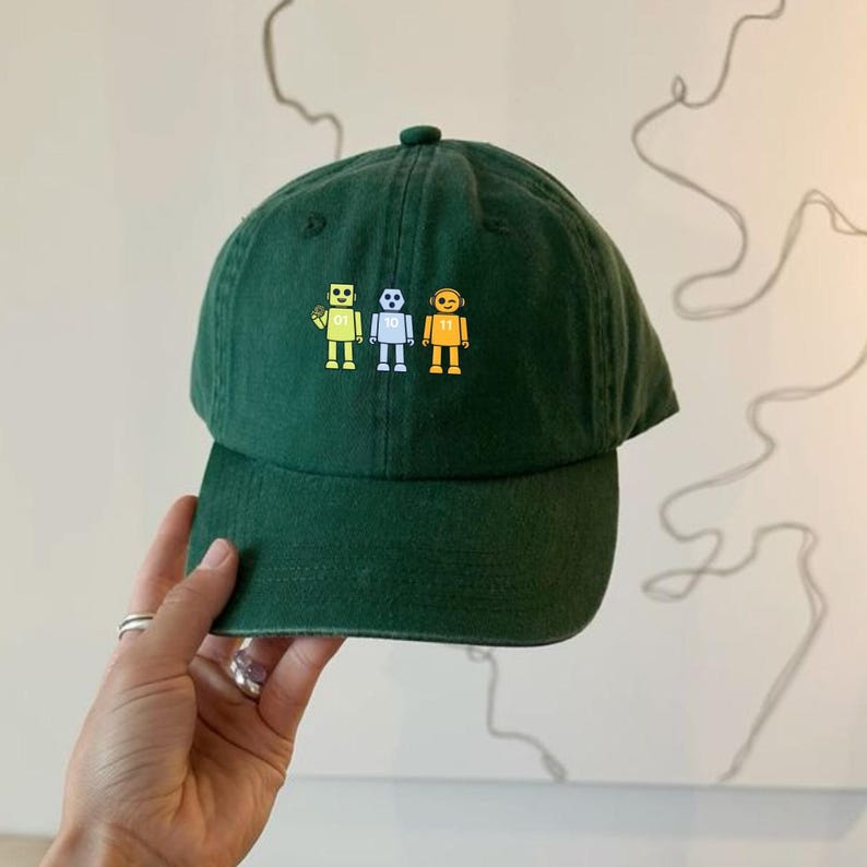 May include: A dark green baseball cap with a curved peak. The cap displays three small robot figures in yellow, white, and orange. The robots have black outlines and are centered on the front. The cap is made of a soft fabric.