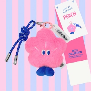 May include: A pink fluffy flower-shaped keychain with blue feet, embroidered eyes, and a blue knotted rope lanyard with silver metal tips. Attached is a white tag labelled "dinhclub" and a product card reading "Fluffy Keychain PEACH TET COLLECTION".