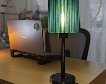 May include: A green, ribbed lampshade sits atop a black lamp base and stem. The lamp is on a wooden desk, with a laptop in the background. The lamp's shade is cylindrical and emits a soft glow.