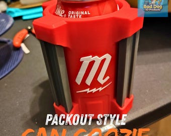 May include: A red can coozie with a white "M" logo and gray accents, holding a Coke can. The coozie has the words "PACKOUT STYLE CAN COOZIE" printed on it. The coozie is designed to keep beverages cold.