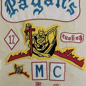 May include: A collection of embroidered patches with the text "Pagan's" in blue gothic lettering. Other patches include "1%", "MC", and a figure holding a cross. The patches are in various colors, including red, yellow, and black.