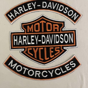 Puede incluir: Parches bordados de Harley-Davidson en negro, naranja y blanco. Los parches incluyen el logotipo de Harley-Davidson y las palabras "MOTORCYCLES" y "CYCLES". Los parches están sobre un fondo beige claro.