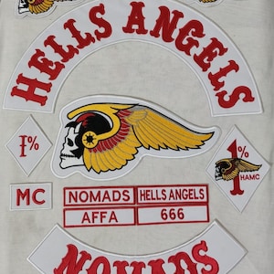 Op de afbeelding: Een verzameling geborduurde patches met de rode woorden "HELLS ANGELS", gebogen over een witte achtergrond. Andere patches bevatten een schedel met vleugels, het woord "NOMADS" en het nummer "666".