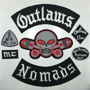 Peut inclure: Ensemble de patchs brodés avec les mots "Outlaws" et "Nomads" en écriture blanche sur fond noir. Un patch central représente un crâne gris avec des yeux rouges et des pistons. D'autres patchs incluent "mc" et un geste de la main.