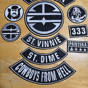 May include: A collection of black and white embroidered patches with various designs and text. The patches include the band name "PANTERA" in a curved banner, a circular logo, and other designs with text such as "ST. VINNIE", "ST. DIME", and "COWBOYS FROM HELL".