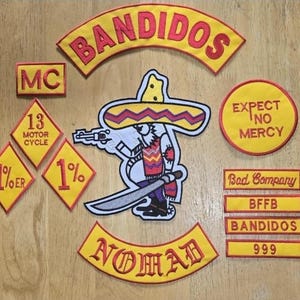 May include: A selection of embroidered patches with a biker theme. The patches display text including "BANDIDOS", "NOMAD", and "EXPECT NO MERCY". A central patch shows a cartoon character with a gun and sword, wearing a sombrero. The patches are yellow and red.