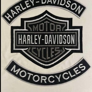 Op de afbeelding: Geborduurde Harley-Davidson patches in zwart en zilver. De patches bevatten het Harley-Davidson logo en de woorden "HARLEY-DAVIDSON" en "MOTORCYCLES" in een gebogen ontwerp. De patches staan op een crèmekleurige achtergrond.
