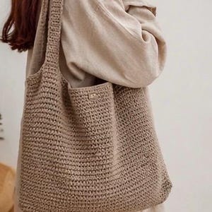 May include: A beige, crocheted tote bag with a shoulder strap. The bag has a large main compartment and a smaller pocket on the front. The crochet pattern is visible, and the bag appears to be made of a natural fibre.