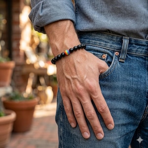 May include: Black beaded bracelet with a rainbow-colored accent bead. The bracelet is worn on a wrist, with the person wearing a blue denim shirt and jeans. The bracelet is a stretch style.