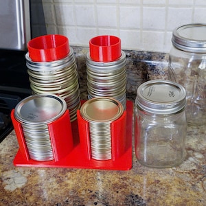 May include: A collection of canning supplies, including clear glass jars with silver lids, stacks of silver jar lids, and red 3D-printed lid holders. The jars are in various sizes, and the lids are neatly organized.