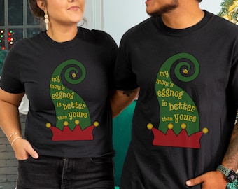 May include: Black short-sleeved t-shirts with a festive design. The shirts feature a green elf hat graphic with the text "my mom's eggnog is better than yours" in gold lettering. A fun holiday theme.