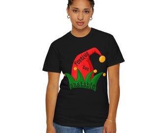 May include: Black t-shirt featuring a festive jester hat design. The hat is red with yellow pom-poms and green accents, with the words "festive but unstable" printed on it. The shirt is worn by a person.