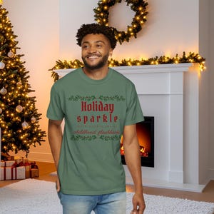 May include: A green t-shirt with the words "Holiday sparkle with nostalgia & childhood flashbacks" in red and white. The shirt is worn by a person in front of a Christmas tree and fireplace.