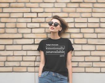 May include: Black t-shirt with the text "Today's forecast: emotionally unavailable again" in white. The shirt is worn with blue jeans and sunglasses. The person is leaning against a brick wall.