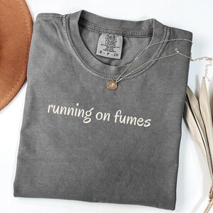 May include: A gray Comfort Colors t-shirt with the phrase "running on fumes" in white script. A gold-colored necklace with a pendant rests on the shirt. A brown hat is visible in the upper left corner.
