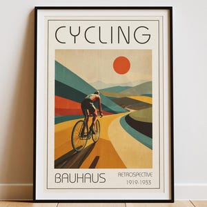 May include: Framed art print with a retro design of a cyclist on a winding road. The word "CYCLING" is at the top, with "BAUHAUS" and "RETROSPECTIVE 1919-1933" at the bottom. The color palette includes orange, yellow, and blue, reminiscent of vintage posters.