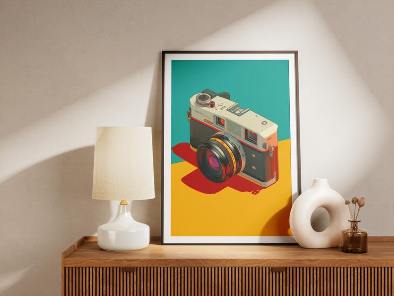 May include: A framed art print featuring a vintage camera illustration. The camera is depicted in a pop art style with a black and silver body, a large lens, and a red shutter button. The background is divided into teal, red, and yellow sections.