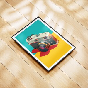 May include: A framed art print featuring a vintage camera illustration. The camera is white and black with a red and black lens, set against a teal and yellow background. The print is in a black frame, resting on a wooden surface.