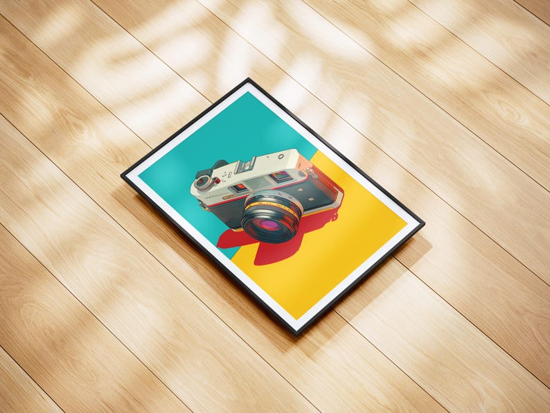 May include: A framed art print featuring a vintage camera illustration. The camera is white and black with a red and black lens, set against a teal and yellow background. The print is in a black frame, resting on a wooden surface.