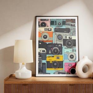 Pu&ograve; includere: Una stampa artistica incorniciata con un collage di fotocamere vintage in vari colori, tra cui rosa, arancione, nero e giallo. Le fotocamere sono disposte su uno sfondo azzurro chiaro, creando un'estetica retr&ograve;. La stampa &egrave; esposta su una superficie di legno.