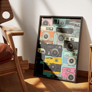 Pu&ograve; includere: Una stampa artistica incorniciata con un collage di fotocamere vintage in vari colori, tra cui rosa, arancione, giallo e nero. Le fotocamere sono disposte su uno sfondo azzurro. La stampa &egrave; esposta accanto a una sedia di legno.