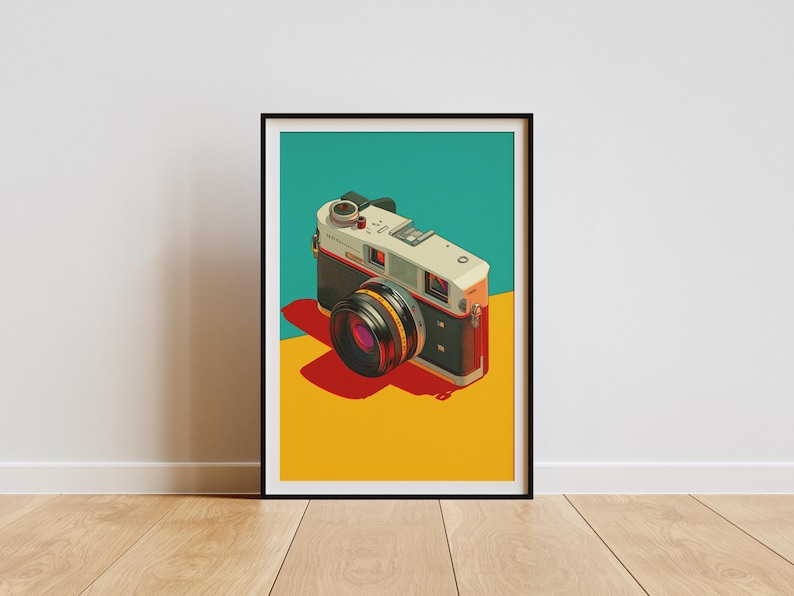 May include: A framed art print showing a vintage camera illustration. The camera has a black and white body, a yellow lens, and a red shadow. The background is split into teal and yellow sections. The print is displayed on a wooden floor.