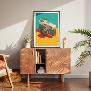 May include: A framed art print featuring a vintage camera illustration. The camera is depicted in a retro style with a teal, yellow, and red color scheme. The print is displayed on a wooden cabinet in a living room setting.