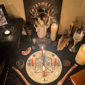 May include: A mystical altar setup featuring a central white plate with a geometric design and lit candles. The scene includes a statue, a skull, a framed picture, and various ritualistic items. The candles are orange and blue.