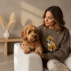 May include: A brown sweatshirt with a dog graphic, worn with cream trousers. A fluffy, light brown dog sits on a white sofa. The dog has curly fur and is looking forward. The background is a neutral colour.