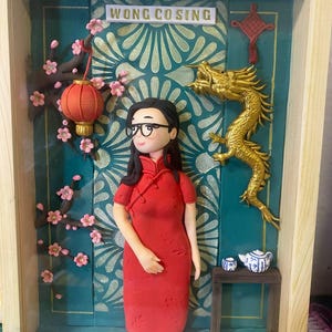 Puede incluir: Diorama enmarcado que presenta a una mujer con un vestido rojo, gafas y un peinado tradicional chino. El fondo incluye un dragón dorado, una linterna y el texto "WONG CO SING". Flores de cerezo y una pequeña mesa con tetera y tazas completan la escena.